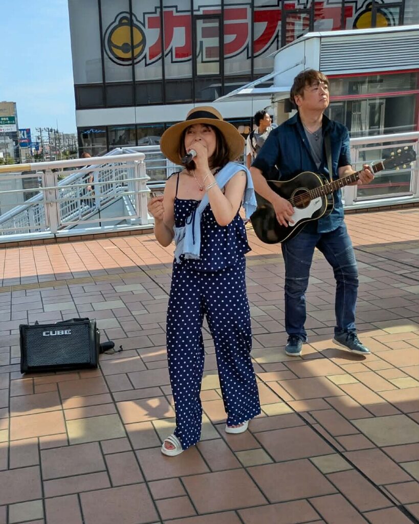 海老名の街並みを背景に演奏する本牧シンガーのんとみっちぃ