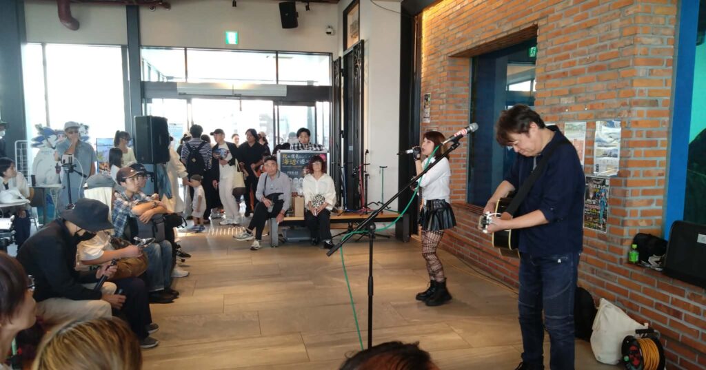 千葉みなと さんばしまつり2026のなかステージで演奏するのんみちと、座って聴くたくさんの観客